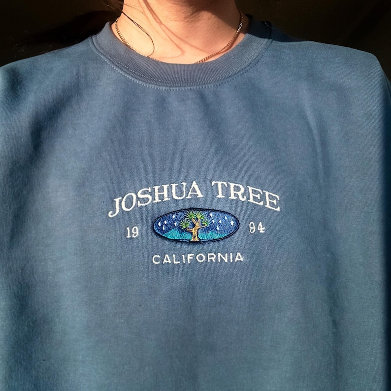 May include: A blue sweatshirt with a white embroidered design that says "Joshua Tree 19 84 California" with a stylized Joshua Tree inside an oval.