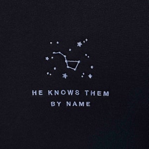 Puede incluir: Camiseta negra con una constelaci&oacute;n bordada en blanco y el texto "HE KNOWS THEM BY NAME".