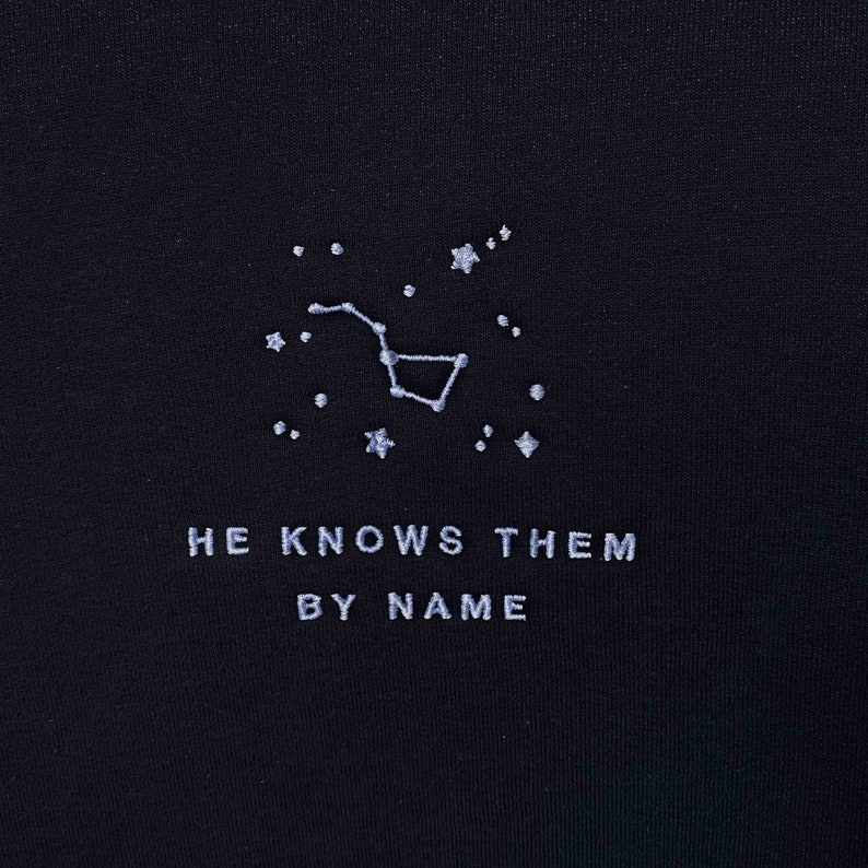 Puede incluir: Camiseta negra con una constelaci&oacute;n bordada en blanco y el texto "HE KNOWS THEM BY NAME".