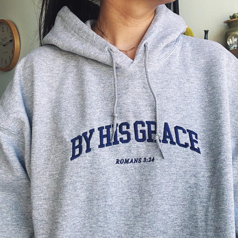May include: Grey hooded sweatshirt with the text "BY HIS GRACE" in blue lettering and "ROMANS 3:24" in smaller lettering below.