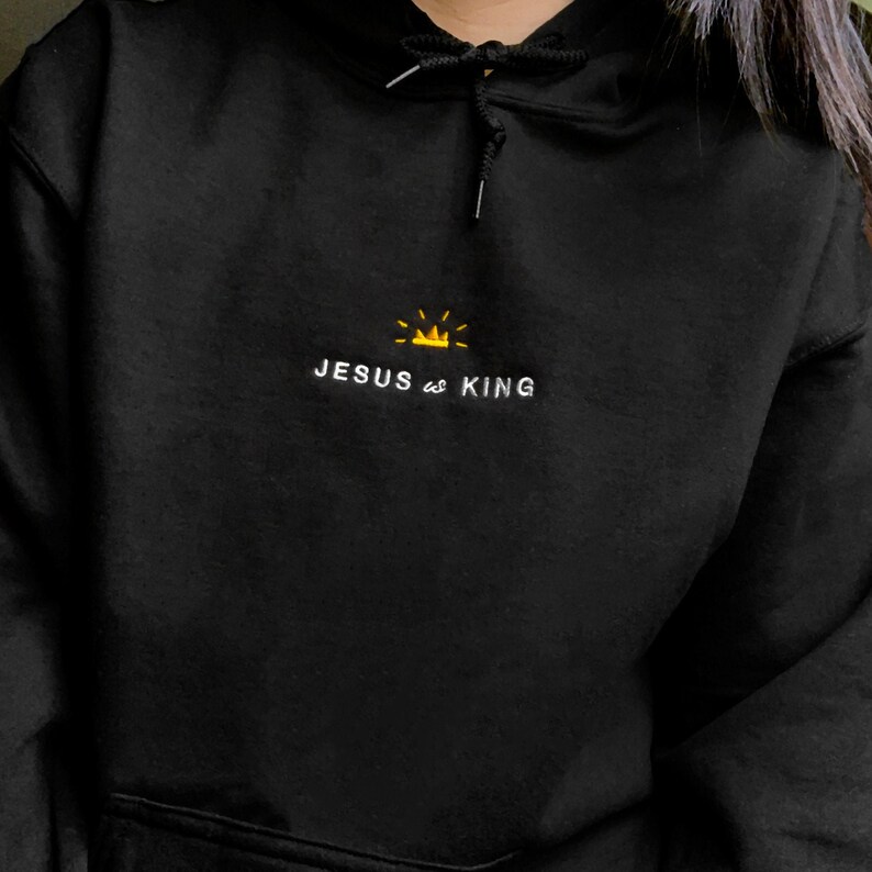 以下が含まれることがあります： 黒いフード付きのスウェットシャツに、白い刺繍で「JESUS is KING」と書いてあり、その上に小さな黄色の王冠が刺繍されています。