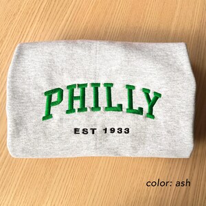 May include: A folded ash-colored sweatshirt with the word "PHILLY" embroidered in green, arched across the front. Below the word is the text "EST 1933" in black. The sweatshirt is made of a soft material.