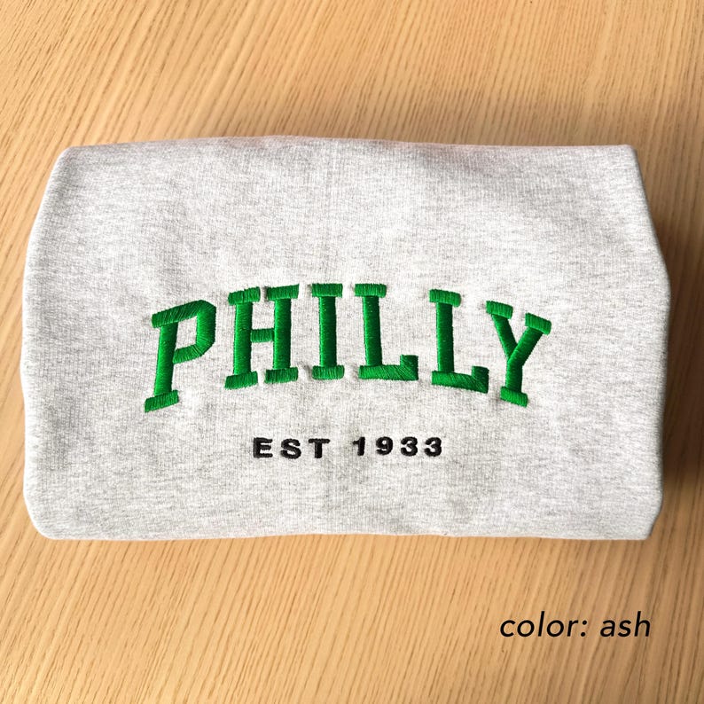 May include: A folded ash-colored sweatshirt with the word "PHILLY" embroidered in green, arched across the front. Below the word is the text "EST 1933" in black. The sweatshirt is made of a soft material.