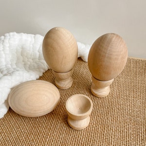 May include: Four wooden eggs, two with stands, one without a stand, and one small wooden egg cup. The eggs are a light brown colour and are on a brown hessian surface.