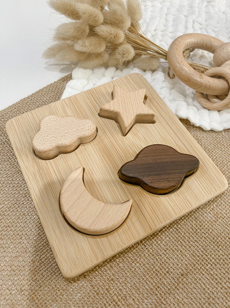 May include: A wooden puzzle with four shapes: a star, a cloud, a crescent moon, and a planet. The puzzle pieces are made of light and dark wood and fit into corresponding cutouts on a bamboo board.