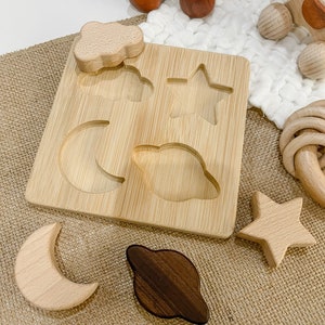 May include: A wooden puzzle board with cutouts of a star, moon, cloud, and planet. The puzzle pieces are made of wood and are scattered around the board. The board is made of bamboo and has a natural finish.