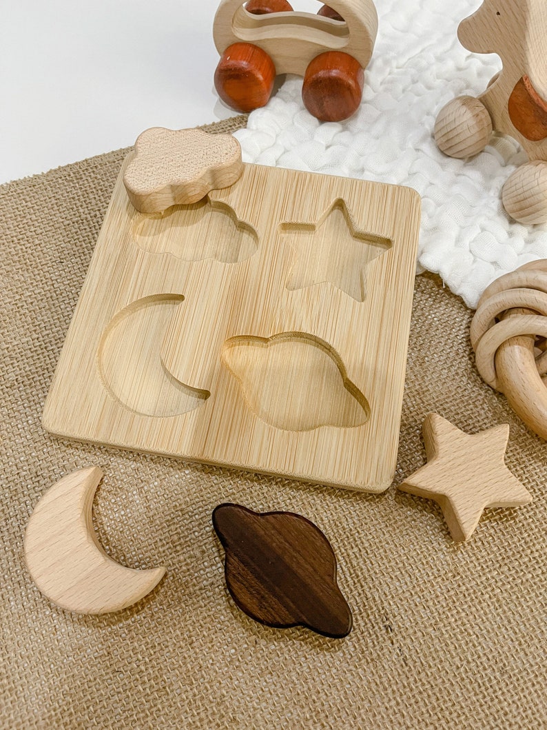 May include: A wooden puzzle board with cutouts of a star, moon, cloud, and planet. The puzzle pieces are made of wood and are scattered around the board. The board is made of bamboo and has a natural finish.