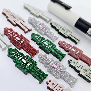 May include: A collection of small, flat nutcracker-shaped charms in various colours including red, green, silver, and white. Each charm features a glittery finish and a small hole at the top for hanging. A white marker is visible in the upper right corner.