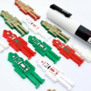 May include: A collection of small, flat nutcracker-shaped charms in red, green, white, and gold colours, arranged alongside a white marker. Each charm features a star detail on the hat and a moustache, with a small loop at the top for hanging.