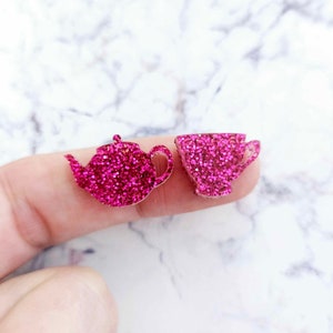 May include: A pair of pink glitter earrings shaped like a teapot and a teacup.