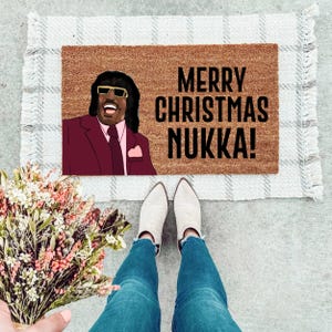 May include: A brown doormat with the text "Merry Christmas Nukka!" in black lettering. A cartoon image of a man with a mustache and sunglasses is on the left side of the doormat.