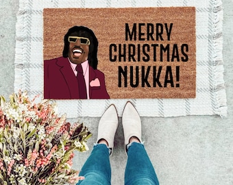 May include: A brown doormat with the text "Merry Christmas Nukka!" in black lettering. A cartoon image of a man with a mustache and sunglasses is on the left side of the doormat.