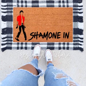 May include: A brown coir doormat with black and white checkered trim and fringe. The doormat has a black silhouette of a person in a red jacket and the text "SHAMONE IN" in black.