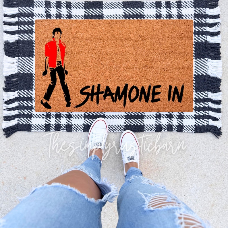 May include: A brown coir doormat with black and white checkered trim and fringe. The doormat has a black silhouette of a person in a red jacket and the text "SHAMONE IN" in black.