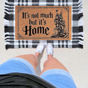 May include: A rectangular tan coir doormat featuring black text that reads It's not much but it's Home, accompanied by a black graphic illustration of a multi-story house with a weather vane, layered over a black and white plaid fabric rug.