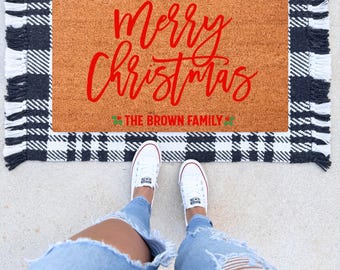 May include: A rectangular doormat with a brown center and a black and white checkered border. The mat features the red text "Merry Christmas" and "THE BROWN FAMILY". White sneakers and blue jeans are visible.
