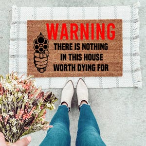May include: A brown doormat with the text "WARNING THERE IS NOTHING IN THIS HOUSE WORTH DYING FOR" in black lettering. A black silhouette of a gun is pointing to the left of the text. The doormat is on a white and gray checkered rug.
