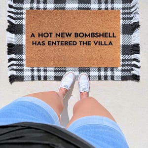 May include: A rectangular tan coir doormat featuring the black text A HOT NEW BOMBSHELL HAS ENTERED THE VILLA, placed on top of a black and white plaid rug with fringed edges. The doormat is positioned on a light-colored surface.