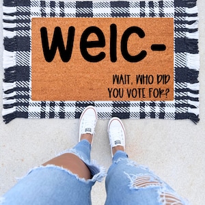 May include: A doormat with the word "welc-" in large black letters, with the phrase "WAIT, WHO DID YOU VOTE FOR?" below. The mat has a brown center and a black and white checkered border. White sneakers and blue jeans are visible.