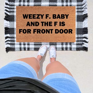 May include: A rectangular tan coir doormat featuring the black text WEEZY F. BABY AND THE F IS FOR FRONT DOOR. The mat is layered over a black and white plaid rug with fringed edges, creating a layered look for an entryway or porch area.