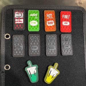 May include: A black fabric patch board with various colorful patches attached. The patches feature cartoon characters, logos, and text. Some of the text includes "Diablo", "Meds", "Hot", "Fire", and "EDC".