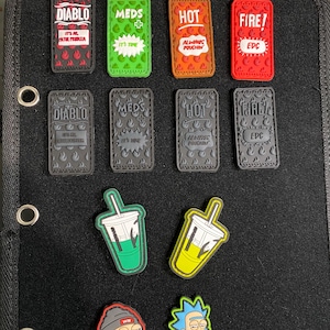 May include: A collection of colorful PVC patches featuring various designs and text. The patches include phrases like "DIABLO," "MEDS," and "FIRE!" along with cartoon drink and character designs. The patches are attached to a black surface.