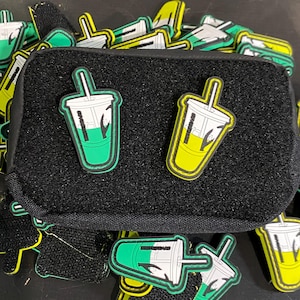 May include: A close-up of a black pouch with two green and yellow drink patches attached. The pouch has a velcro closure and is surrounded by a pile of similar patches.