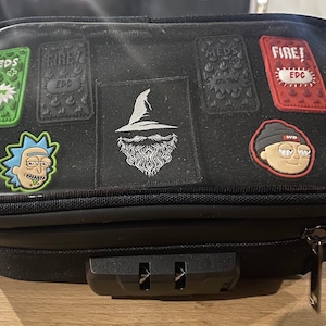 May include: Black zippered pouch with a clear top section, featuring multiple patches. Patches include a wizard, cartoon characters, and text that reads "MEDS", "FIRE! EDC". The pouch has a combination lock and a zipper closure.