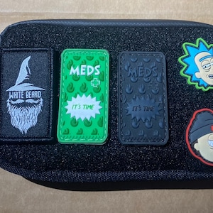 May include: Black rectangular pouch with multiple patches. Patches include a white wizard with the text "WHITE BEARD", two "MEDS IT'S TIME" patches, and two cartoon character patches. The pouch has a loop on the side.
