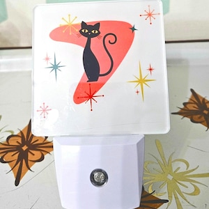 Könnte beinhalten: Ein weißes Nachtlicht mit Retro-Design, das eine schwarze Katzen-Silhouette auf einer roten Bumerangform zeigt. Das Design umfasst Sternausbrüche in Gelb, Rot und Blau. Das Nachtlicht hat einen weißen Sockel mit einem kleinen, runden Licht.