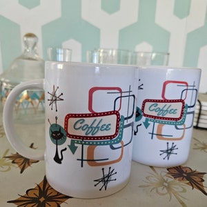 May include: Two white ceramic mugs with a retro design featuring a black cat, geometric shapes, and the word "Coffee" in a blue and red sign. The mugs are on a table with a floral pattern.