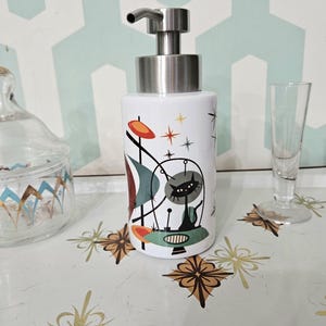 Puede incluir: Dispensador de jabón de cerámica blanca con bomba de acero inoxidable. El dispensador presenta un diseño retro con un gato negro, estrellas y formas geométricas en naranja, verde azulado y marrón. Sobre una superficie blanca con estampado floral.