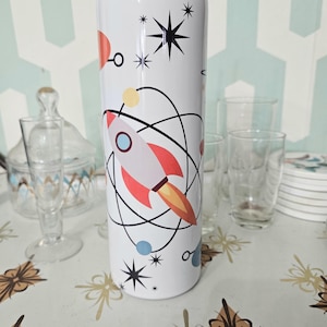 May include: White stainless steel tumbler with a rocket ship, planets, and stars design. The tumbler has a lid and a straw.