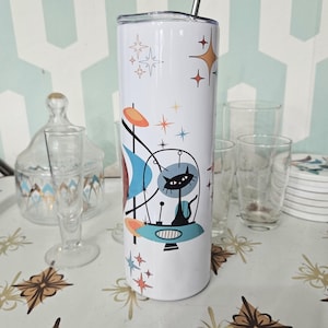 Puede incluir: Un vaso de acero inoxidable blanco con un diseño retro que presenta un gato negro en una nave espacial con detalles coloridos. El vaso tiene una pajita y está sobre una mesa con un mantel estampado.