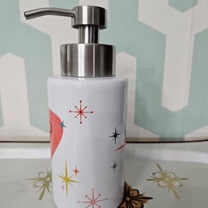 Puede incluir: Dispensador de jab&oacute;n de cer&aacute;mica blanca con bomba de acero inoxidable. El dispensador est&aacute; decorado con un coraz&oacute;n rojo y dise&ntilde;os de explosiones de estrellas de colores. El dispensador es cil&iacute;ndrico y se encuentra sobre una bandeja decorativa.