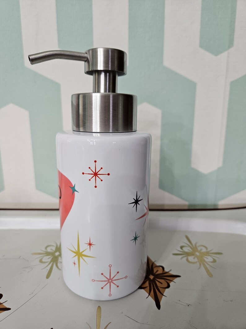 Puede incluir: Dispensador de jab&oacute;n de cer&aacute;mica blanca con bomba de acero inoxidable. El dispensador est&aacute; decorado con un coraz&oacute;n rojo y dise&ntilde;os de explosiones de estrellas de colores. El dispensador es cil&iacute;ndrico y se encuentra sobre una bandeja decorativa.