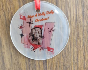May include: A round, clear, glass ornament with a red ribbon. The ornament has a picture of Dolly Parton with the text "Have A Holly Dolly Christmas!" printed on it.