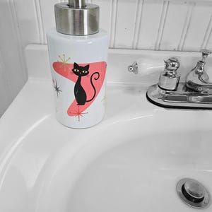 Puede incluir: Dispensador de jab&oacute;n de cer&aacute;mica blanca con un dise&ntilde;o de gato negro. El dispensador tiene una bomba de acero inoxidable y un dise&ntilde;o at&oacute;mico rosa y dorado de estilo retro. El gato tiene ojos amarillos y una cola curvada.