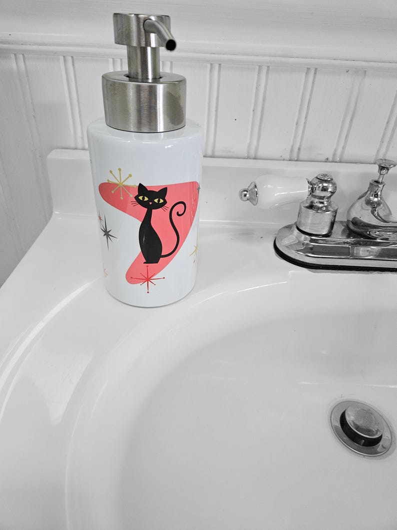 Puede incluir: Dispensador de jab&oacute;n de cer&aacute;mica blanca con un dise&ntilde;o de gato negro. El dispensador tiene una bomba de acero inoxidable y un dise&ntilde;o at&oacute;mico rosa y dorado de estilo retro. El gato tiene ojos amarillos y una cola curvada.