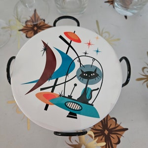 May include: A white ceramic coaster with a black cat in a spaceship design. The coaster has a black metal frame and features abstract shapes in blue, red, orange, and brown.