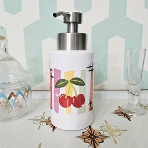 May include: A white ceramic soap dispenser with a stainless steel pump. The dispenser features a retro design with red cherries, green leaves, and colorful stripes. The dispenser is on a patterned surface.