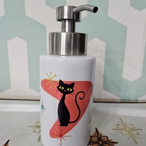 Puede incluir: Dispensador de jab&oacute;n de cer&aacute;mica blanca con bomba de acero inoxidable. El dispensador presenta una silueta de gato negro sobre una forma roja de la era at&oacute;mica, con detalles de estrellas. El dispensador es cil&iacute;ndrico y tiene un dise&ntilde;o moderno y retro.