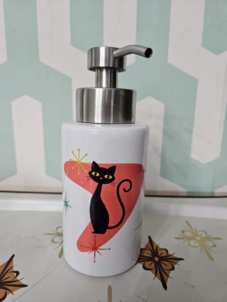 Puede incluir: Dispensador de jab&oacute;n de cer&aacute;mica blanca con bomba de acero inoxidable. El dispensador presenta una silueta de gato negro sobre una forma roja de la era at&oacute;mica, con detalles de estrellas. El dispensador es cil&iacute;ndrico y tiene un dise&ntilde;o moderno y retro.