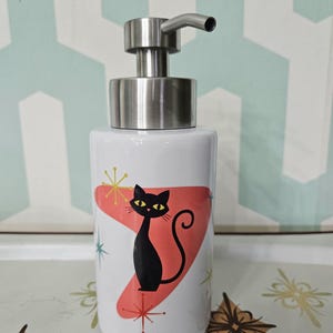 Puede incluir: Dispensador de jab&oacute;n de cer&aacute;mica blanca con bomba de acero inoxidable. El dispensador presenta una silueta de gato negro sobre una forma de boomerang roja, con detalles de estrellas. Un accesorio de ba&ntilde;o de estilo retro.