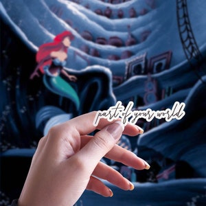 May include: A white sticker with the text "part of your world" in a cursive font. The sticker is held in front of a blurred background featuring a mermaid with red hair and a blue underwater scene.