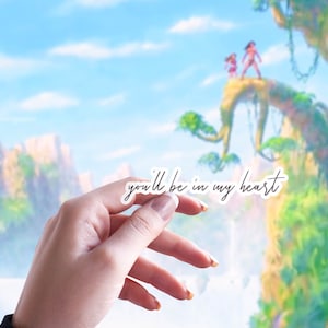May include: A hand holding a white sticker with the text "you'll be in my heart" in black script. The sticker is in front of a blurred background of a blue sky, green trees, and a waterfall.