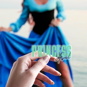 Op de afbeelding: Een transparante sticker met de woorden "PRINCESS Ariel" in groen en zwart. De sticker wordt vastgehouden voor een persoon die een blauwe jurk en een zwarte top draagt.
