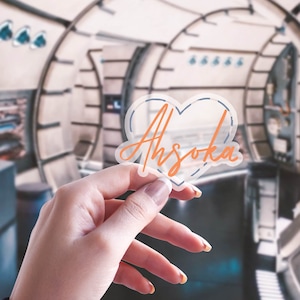 K&ouml;nnte beinhalten: Ein klarer herzf&ouml;rmiger Aufkleber mit dem Namen "Ahsoka" in orangefarbener Schrift. Der Aufkleber hat eine blaue Umrandung und wird vor einem verschwommenen Hintergrund gehalten. Der Aufkleber ist etwa 7.6 cm breit.