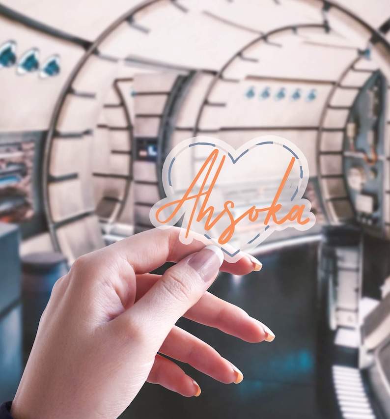 K&ouml;nnte beinhalten: Ein klarer herzf&ouml;rmiger Aufkleber mit dem Namen "Ahsoka" in orangefarbener Schrift. Der Aufkleber hat eine blaue Umrandung und wird vor einem verschwommenen Hintergrund gehalten. Der Aufkleber ist etwa 7.6 cm breit.