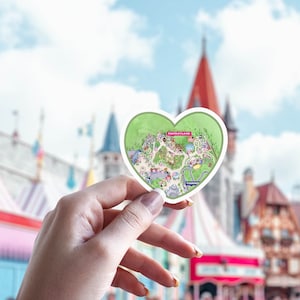 May include: A heart-shaped sticker featuring a map of Fantasyland. The map is green with colorful illustrations of various attractions. The sticker is held in front of a blurred background of a theme park with blue skies.